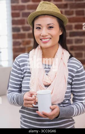 Femme asiatique adulte moyenne debout dans le salon portant Fedora en paille tenant une tasse en céramique blanche et souriante Banque D'Images