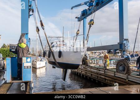 Warns, pays-Bas - 20 novembre 2023 : grue de levage de bateau hissant un voilier hors de l'eau dans une marina animée, avec des ouvriers habilement opératinés Banque D'Images