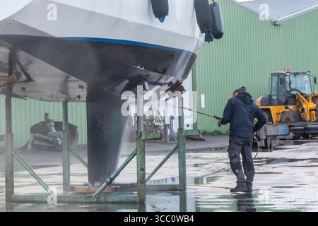 Warns, pays-Bas - 20 novembre 2023 : un ouvrier de chantier naval nettoie la coque d'un voilier avec un nettoyeur haute pression, alors qu'il est soulevé par une grue, entouré Banque D'Images
