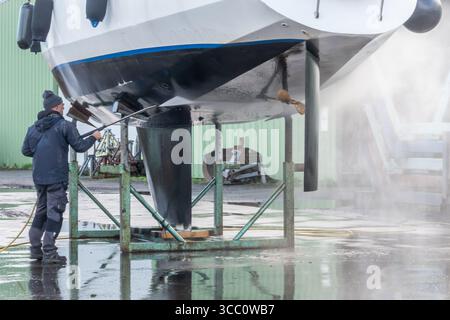 Warns, pays-Bas - 20 novembre 2023 : un ouvrier de chantier naval nettoie la coque d'un voilier avec un nettoyeur haute pression, alors qu'il est soulevé par une grue, entouré Banque D'Images