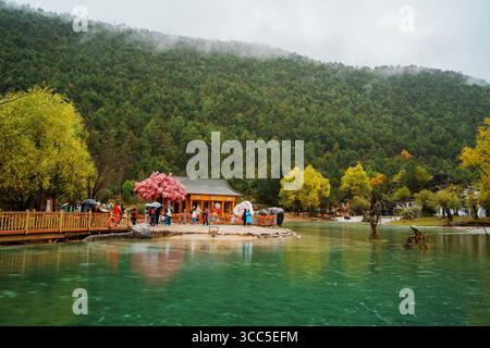 Lijiang, Chine - 24 octobre 2024 : Pavillon traditionnel en bois avec des fleurs roses près d'un lac turquoise dans la vallée de la lune bleue, entouré d'une montagne verdoyante Banque D'Images