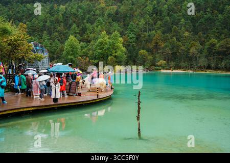 Lijiang, Chine - 24 octobre 2024 : les visiteurs avec des parasols apprécient les eaux turquoises de Blue Moon Valley, avec des collines boisées luxuriantes en arrière-plan. Banque D'Images