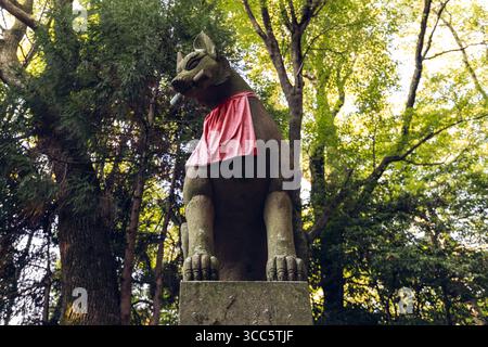 Statue de renard en pierre avec une clé dans la bouche et un bavoir rouge au sanctuaire de Fushimi Inari à Kyoto, Japon, entourée d'une forêt sacrée et d'une lumière du jour doucement filtrée Banque D'Images
