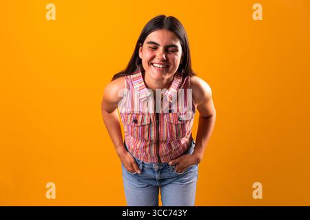 Jeune femme joyeuse aux longs cheveux foncés, portant un haut rayé coloré et un Jean, souriant les yeux fermés tout en se penchant en avant contre un o vibrant Banque D'Images