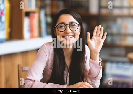 Appel vidéo. Heureuse jeune femme arabe saluant quelqu'un, ayant une conférence virtuelle, agitant de la main à la caméra et souriant Banque D'Images