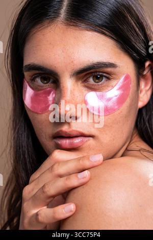 Belle jeune femme avec des taches de rousseur reçoit un traitement de soin de la peau, en appliquant des patchs cosmétiques roses sous ses yeux, favorisant le bien-être, la beauté et le se Banque D'Images