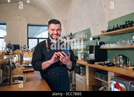 Propriétaire de petite entreprise souriant vêtu d'un uniforme de chef noir avec un tablier à l'aide d'un smartphone dans sa propre salle de restaurant confortable. Les gens qui réussissent, dur w Banque D'Images