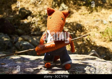 Un renard jouet tricoté tenant une saucisse rôtie sur un bâton, avec un fond sauvage. Photographié en juillet 2025. Banque D'Images