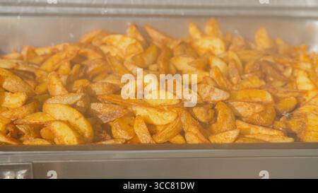 Gros plan des quartiers de pommes de terre assaisonnés dans un buffet chaud. Chaud, croustillant et doré, parfait pour la restauration, les menus de mariage, les promotions de restaurant, blogs alimentaires Banque D'Images