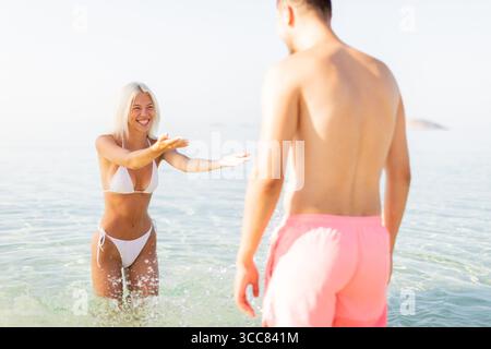 Une jeune femme portant un bikini salue joyeusement un ami dans l'eau de l'océan peu profonde. Le soleil brille, créant une atmosphère animée remplie de Laug Banque D'Images