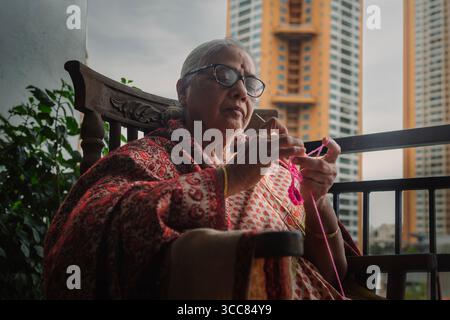 Femme indienne âgée assise sur une chaise à bascule en bois sur un balcon, tricotant avec des boules de fil colorées, portant un saree traditionnel et des lunettes. Banque D'Images