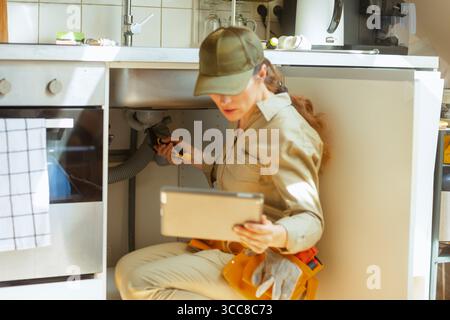 Une femme plombière s'agenouille sous un évier de cuisine avec une clé et une tablette, inspectant un blocage de tuyau et combinant le travail pratique avec des outils numériques. Banque D'Images