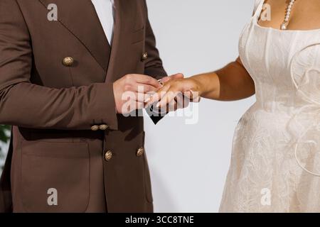 Gros plan d'une cérémonie de mariage capturant un marié glissant délicatement une bague sur le doigt de la mariée. élégant costume marron, robe en dentelle blanche, intimat Banque D'Images