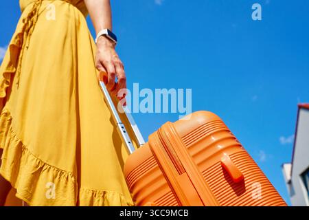 Femme en robe jaune tenant une valise orange contre le ciel bleu avec des nuages. Gros plan d'une voyageuse avec des bagages. Concept de voyage élégant, va Banque D'Images