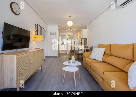 Salon lumineux avec canapé moutarde, téléviseur mural et console multimédia en bois. Banque D'Images