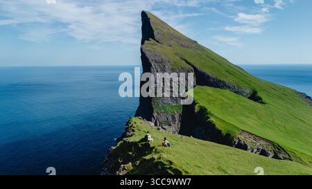 Les aventuriers se prélassent de la beauté époustouflante des îles Féroé, assis au sommet de falaises spectaculaires. Le paysage verdoyant et le vaste océan créent une toile de fond impressionnante sous un ciel bleu clair. Banque D'Images