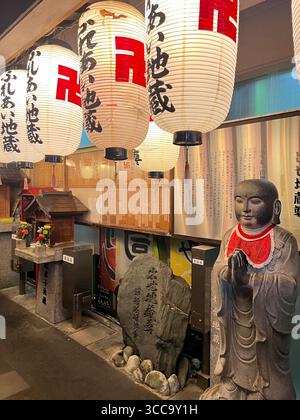 Statue en pierre et sanctuaire avec des lanternes de papier japonaises dans une ruelle chaleureusement éclairée, Japon. Banque D'Images