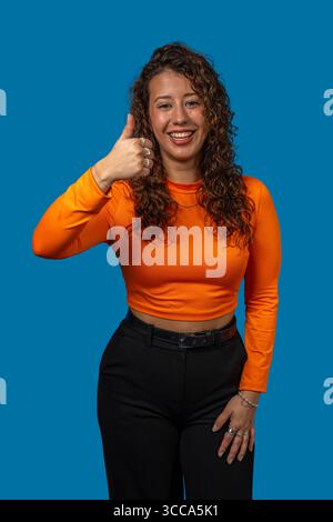 Portrait studio d'une jeune femme joyeuse faisant des gestes pouces vers le haut, véhiculant l'approbation et l'attitude positive sur fond bleu vif Banque D'Images