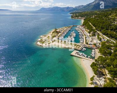 Vue aérienne d'un port de plaisance dynamique où les eaux turquoises rencontrent le rivage sablonneux, contrastant avec les collines verdoyantes et les montagnes lointaines, Krvavica, comté de Split-Dalmatie, Croatie. Banque D'Images