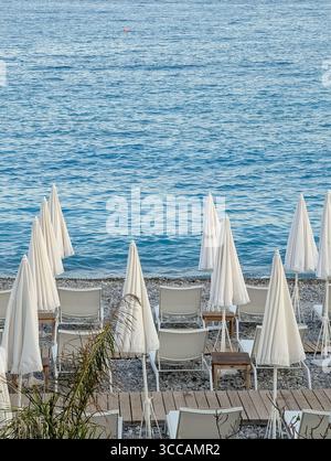Scène matinale tranquille sur l'emblématique plage de galets de Nice, Côte d'Azur, avec des parasols blancs fermés et des chaises longues vides. Banque D'Images