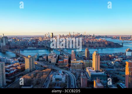 Vue aérienne de la ville et des ponts au-dessus de l'eau, gratte-ciel atteignant le ciel depuis Manhattan, le tout illuminé par la lueur chaude du soleil couchant, New York, États-Unis. Banque D'Images