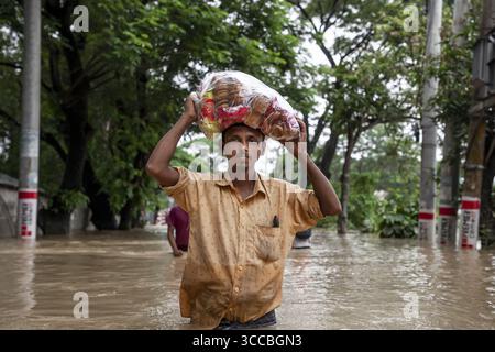 Satkania, Bangladesh - 09 août 2023 : vue d'un homme pataugant dans les eaux de crue, transportant un grand sac d'objets colorés au-dessus de lui, au milieu de rues et d'arbres submergés. Banque D'Images
