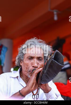 Chandanath hill, Bangladesh - 12 March 2021: View of an elder man with weathered skin blowing into a traditional horn against a vibrant orange backdrop. Banque D'Images