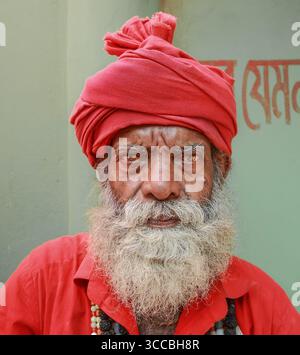 Chandanath hill, Bangladesh - 12 March 2021: View of an elderly man with a striking red turban and weathered face, his intense gaze softened by a long Banque D'Images