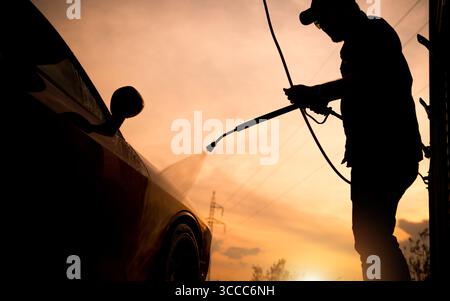 Un homme rince une voiture avec un nettoyeur haute pression au coucher du soleil, projetant une silhouette frappante contre le ciel coloré et soulignant la sérénité du soir. Banque D'Images