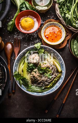 Bol de nouilles asiatiques avec des légumes verts, chou chinois et boulettes de viande sur fond sombre avec des couverts en bois, des baguettes et de sauce chaude, vue d'en haut. Close up Banque D'Images