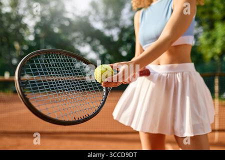 Vue rapprochée d'une joueuse de tennis qui se prépare à servir sur un court ensoleillé. Concentrez-vous sur sa main avec le ballon et la raquette, montrant l'énergie, l'équilibre, Banque D'Images