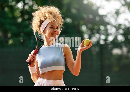 Portrait de femme heureuse avec raquette de tennis et balle sur terrain d'argile, profitant de la pratique estivale. Concept : mode de vie actif, joie du sport, fitness inspirat Banque D'Images