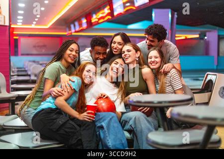 Groupe de jeunes personnes multi-ethniques profitant du temps ensemble, serrant dans leurs bras et riant, assis sur des sièges dans une piste de bowling Banque D'Images