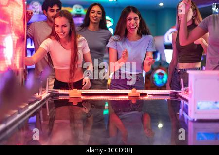 Groupe de jeunes amis jouissant d'un match compétitif de hockey pneumatique dans une arcade vibrante, riant et se encourageant les uns les autres Banque D'Images