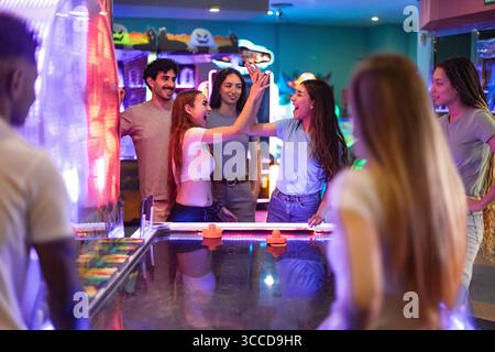 Groupe d'amis heureux jouant au hockey pneumatique et donnant high cinq, célébrant un but dans une arcade d'amusement colorée Banque D'Images