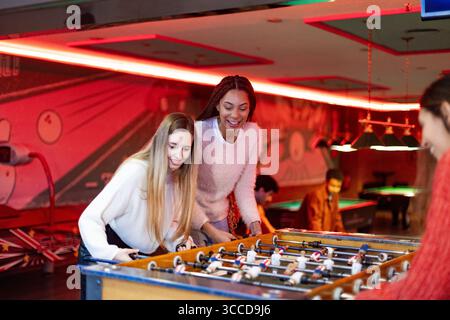 Deux jeunes femmes riant et élaborant des stratégies tout en profitant d'un jeu passionnant de baby-foot dans une salle de jeux animée et colorée Banque D'Images