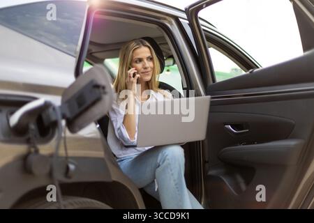 Femme d'affaires assise dans sa voiture électrique pendant qu'elle est en charge, travaillant sur son ordinateur portable et parlant au téléphone Banque D'Images