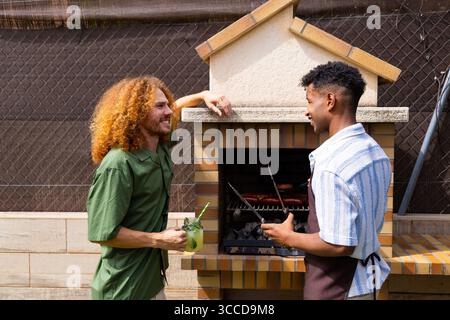 Deux amis mâles grillent des saucisses sur un barbecue dans une cour arrière, profitant d'une journée d'été avec des boissons et des rires Banque D'Images