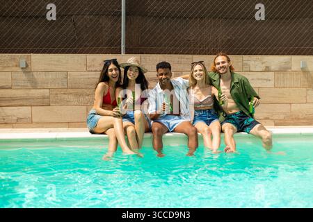 Groupe d'amis multi-ethniques profitant de moments ensoleillés au bord de la piscine, riant et partageant des boissons tout en célébrant leurs vacances insouciantes Banque D'Images