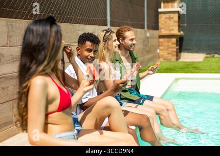 Groupe d'amis divers appréciant des tranches de pastèque rafraîchissantes tout en se relaxant au bord de la piscine par une journée d'été ensoleillée Banque D'Images