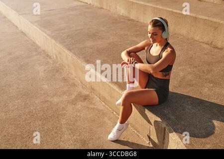 Une jeune femme est assise confortablement sur un ensemble de marches en béton, portant des écouteurs, entièrement absorbée par sa musique préférée, entourée d'un an paisible Banque D'Images