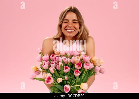 Une jeune femme ravie dans un haut rose rit les yeux fermés, embrassant un grand bouquet de tulipes roses, debout sur un fond rose pastel. Pure Banque D'Images