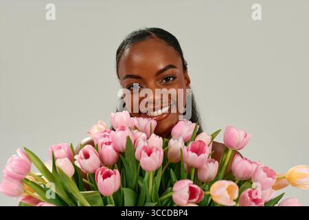 Femme afro-américaine tenant un bouquet de tulipes roses, célébrant la fête des mères ou la journée des femmes avec joie et amour, souriant brillamment sur fond neutre. Conc Banque D'Images