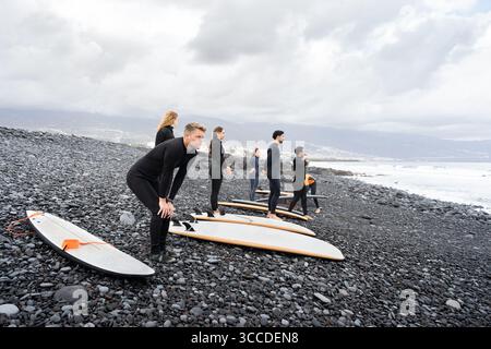 Les élèves de surf portant des combinaisons se réchauffent sur une plage volcanique noire avant une leçon de surf Banque D'Images