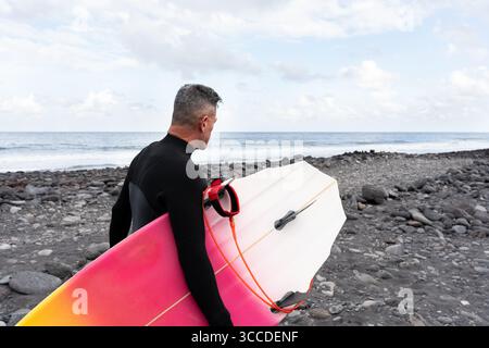 Surfeur mature portant une planche de surf sur une plage rocheuse regardant les vagues de l'océan Banque D'Images