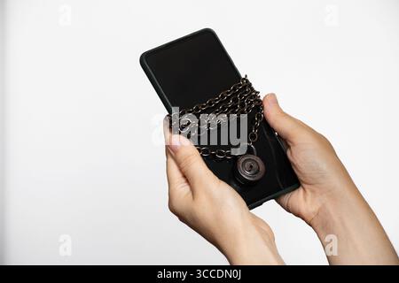 Mains féminines tenant un smartphone enveloppé dans une chaîne métallique avec un cadenas sur fond blanc sur le thème de la sécurité des mots de passe et des données Banque D'Images