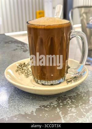 Un café de grailam traditionnel chaud mousseux dans une tasse en verre transparent avec cuillère, sur une soucoupe décorative dans un café rustique kopitiam kuala lumpur malaisie Banque D'Images