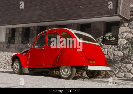Une charmante voiture rouge est garée contre un bâtiment rustique en pierre, avec un ciel bleu clair au-dessus. Cette scène nostalgique capture l'essence d'un paisible Banque D'Images
