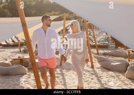 Un couple se promène main dans la main le long d'une plage ensoleillée, entourée de verrières élégantes et de salons. Banque D'Images