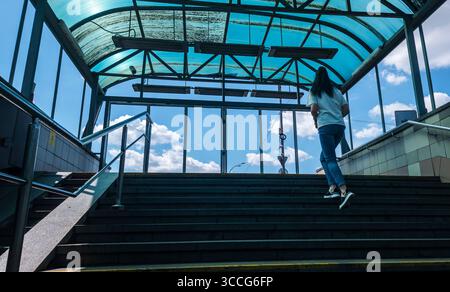 Kiev, Ukraine - 25 juillet 2025 : jeune femme montant les escaliers dans une station de métro de Kiev, sous un toit bleu transparent, avec un ciel bleu vif et blanc c Banque D'Images